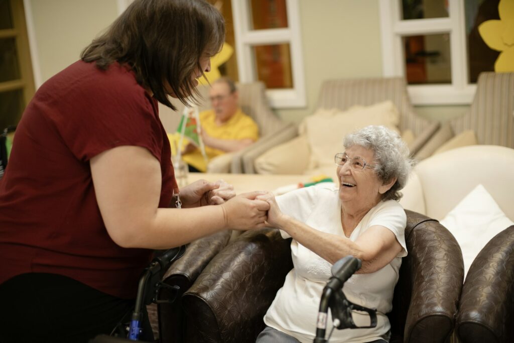 A caregiver holds the hands of a smiling older woman seated with her walker